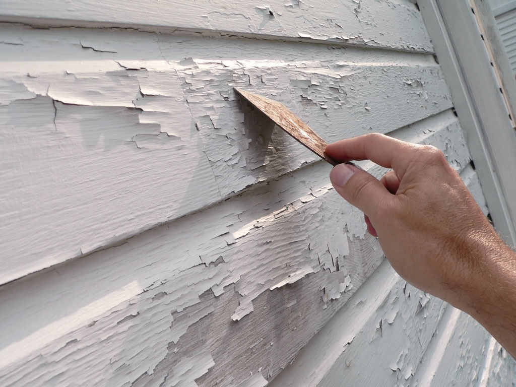 Image of a hand using a paint scraper to scrape off the old exterior paint of a home